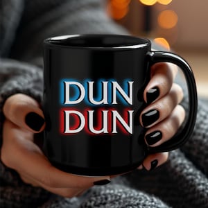 May include: Black ceramic mug with the text "DUN DUN" in white, with blue and red neon-style outlines. The mug is held by a person with black nail polish.