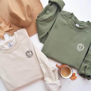 May include: Two crewneck sweatshirts, one beige and one olive green, each with a heart-shaped graphic with flames. A brown hat and a cup of coffee with a leaf are also in the image. The sweatshirts are made of a soft material.