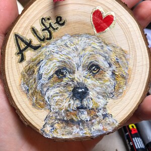 May include: A wooden round ornament with a painted portrait of a white dog with brown markings. The dog's name, "Alfie", is written in black cursive lettering on the ornament. A red heart is painted on the ornament.