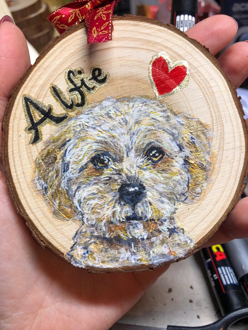 May include: A wooden round ornament with a painted portrait of a white dog with brown markings. The dog's name, "Alfie", is written in black cursive lettering on the ornament. A red heart is painted on the ornament.