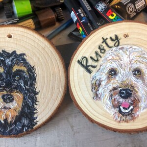 May include: Two wooden round ornaments with painted portraits of dogs. The ornament on the left has a black and brown dog with the name "Flo" painted on it. The ornament on the right has a white and brown dog with the name "Rusty" painted on it.