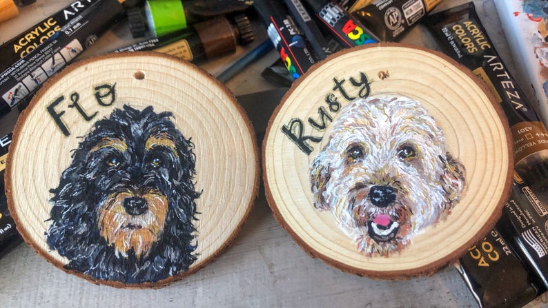 May include: Two wooden round ornaments with painted portraits of dogs. The ornament on the left has a black and brown dog with the name "Flo" painted on it. The ornament on the right has a white and brown dog with the name "Rusty" painted on it.