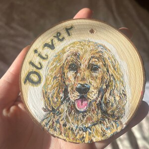 May include: A wooden round ornament with a painted portrait of a golden dog with the name "Oliver" written on the top.
