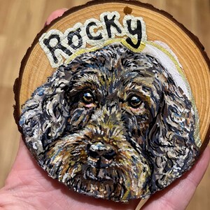 May include: A hand-painted wooden slice with a portrait of a brown and black dog with a halo. The dog's name, "Rocky", is written in gold lettering above the dog.