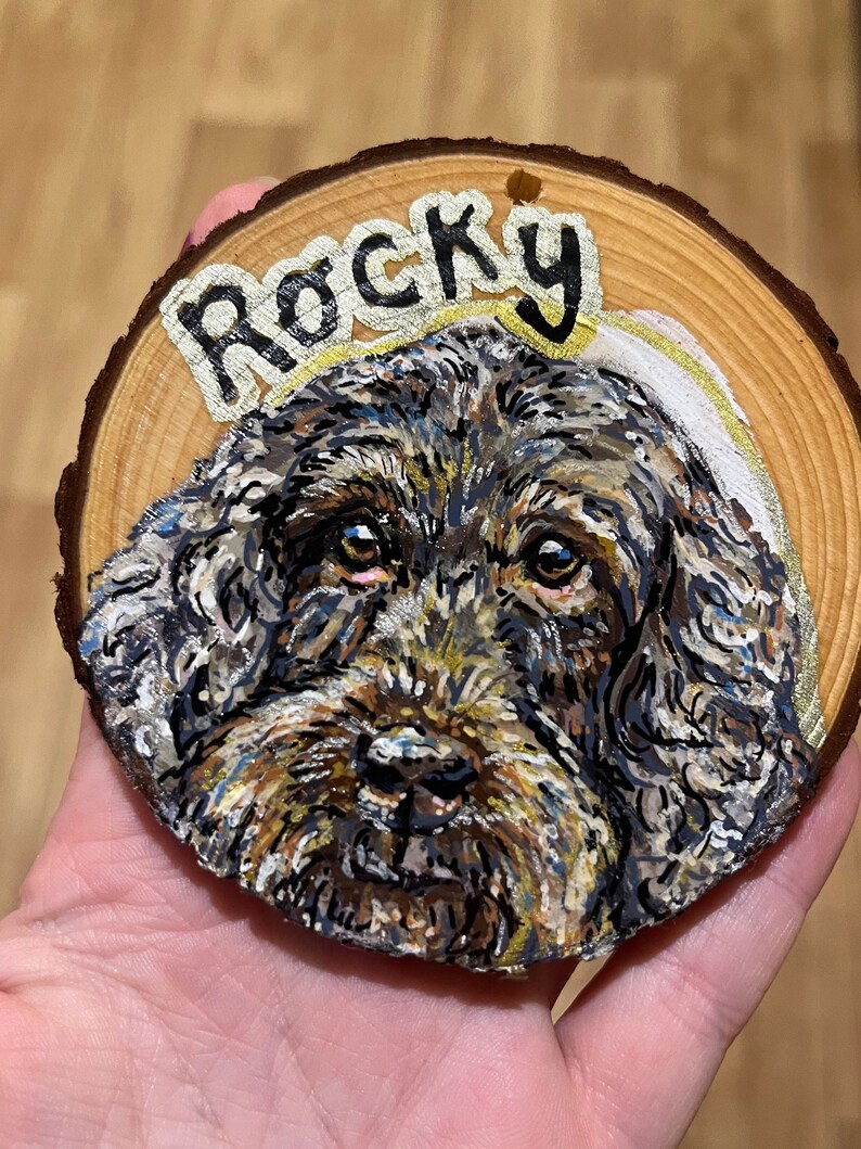 May include: A hand-painted wooden slice with a portrait of a brown and black dog with a halo. The dog's name, "Rocky", is written in gold lettering above the dog.