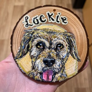 May include: A wooden round ornament with a painted portrait of a dog with the name "Lockie" written above the dog.
