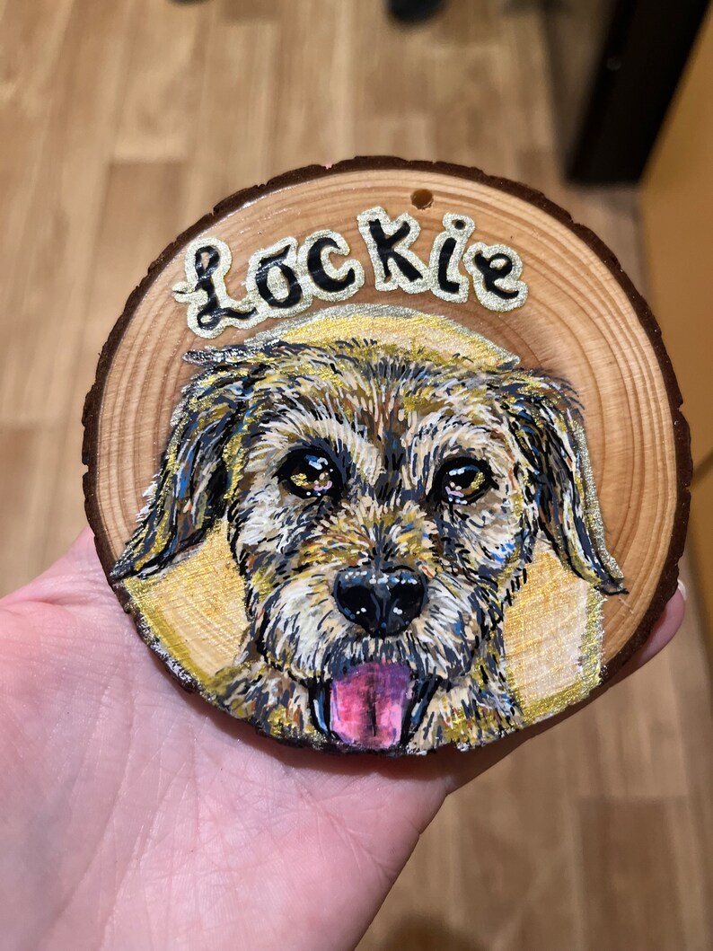 May include: A wooden round ornament with a painted portrait of a dog with the name "Lockie" written above the dog.