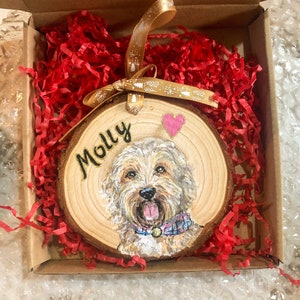 May include: A wooden round ornament with a painted portrait of a dog with the name "Molly" written in gold lettering. The ornament is in a red shredded paper-filled box with a gold ribbon.