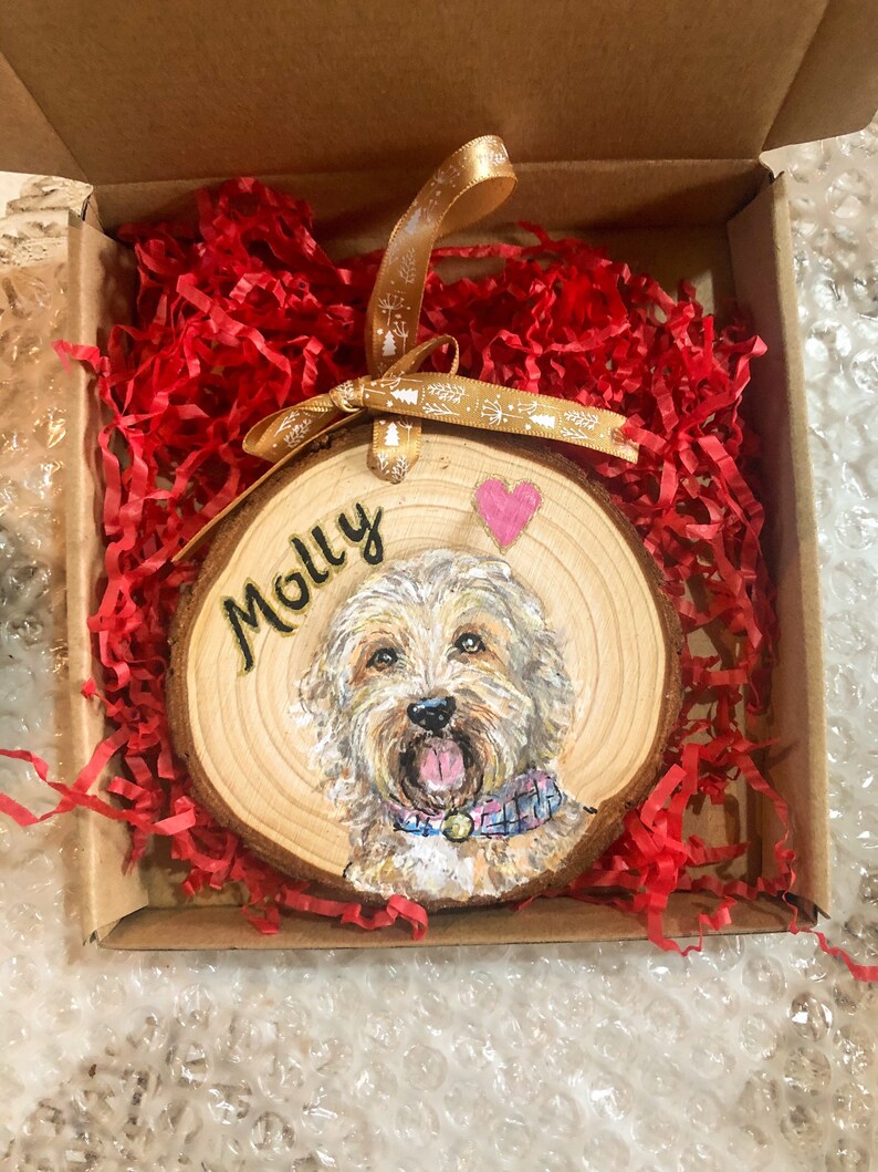 May include: A wooden round ornament with a painted portrait of a dog with the name "Molly" written in gold lettering. The ornament is in a red shredded paper-filled box with a gold ribbon.