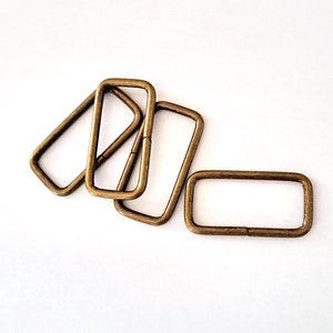 Peut inclure: Cinq anneaux en D en métal de couleur or antique, également appelés boucles de ceinture, sont disposés en pile sur un fond blanc.
