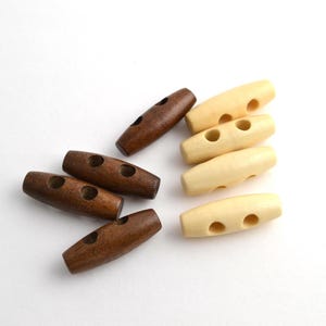 Può includere: Un set di bottoni a levetta in legno, alcuni di colore marrone chiaro e altri di colore marrone scuro. I bottoni hanno due fori ciascuno e sono di forma ovale.