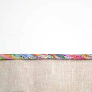 May include: A strip of fabric featuring a vibrant, multi-coloured abstract pattern with streaks of pink, orange, blue, green, and yellow, attached to a solid, light beige, textured linen-like fabric with a visible woven grid pattern.