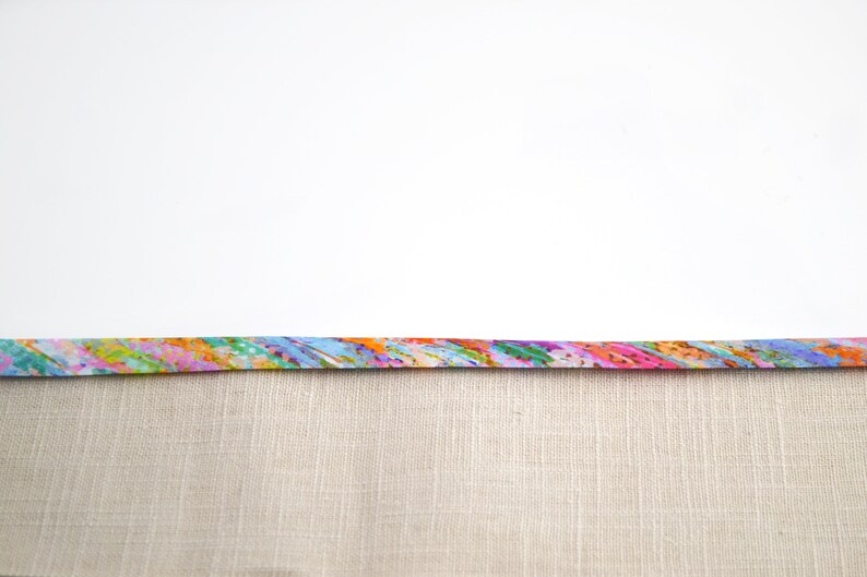 May include: A strip of fabric featuring a vibrant, multi-coloured abstract pattern with streaks of pink, orange, blue, green, and yellow, attached to a solid, light beige, textured linen-like fabric with a visible woven grid pattern.