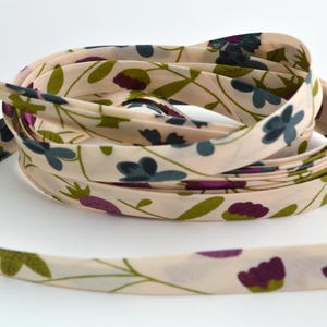 Könnte beinhalten: Eine Rolle cremefarbenes Stoffband mit einem Blumendruck in Grün-, Lila- und Blautönen. Das Band ist gefaltet und aufgerollt.