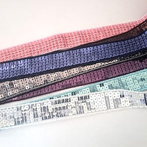 Peut inclure: Gros plan sur cinq galons différents de tissu pailleté. Les galons sont dans des tons de rose, violet, bleu et blanc. Les paillettes sont petites et serrées.