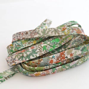 Könnte beinhalten: Eine aufgerollte Länge eines floral gemusterten Schrägbandes. Der Stoff hat einen cremefarbenen Hintergrund mit einem sich wiederholenden Muster aus grünen Blättern, orangefarbenen Blüten und braunen Akzenten. Das Band wird wahrscheinlich zum Nähen oder Basteln verwendet.