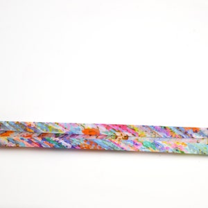 May include: A long, thin, rectangular strip of paper featuring a vibrant, multi-coloured abstract pattern. The design consists of small, dense brushstrokes in shades of orange, pink, blue, green, and yellow, creating a textured, painterly appearance.