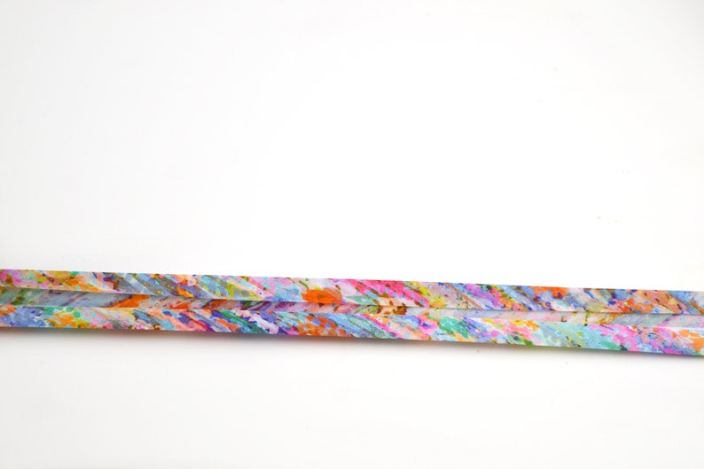 May include: A long, thin, rectangular strip of paper featuring a vibrant, multi-coloured abstract pattern. The design consists of small, dense brushstrokes in shades of orange, pink, blue, green, and yellow, creating a textured, painterly appearance.