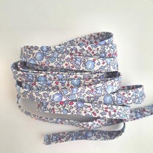 Peut inclure: Un rouleau de garniture en tissu floral avec un motif répété de petites fleurs bleues, violettes et roses sur fond blanc. La garniture est pliée et empilée, mettant en valeur le motif floral.