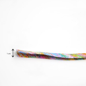 May include: A long, thin strip of paper featuring a vibrant, multi-coloured abstract pattern with splashes of pink, blue, yellow, and green. A black arrow graphic indicates a width of 1 cm on the left side of the strip against a plain white background.