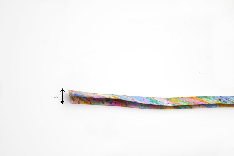 May include: A long, thin strip of paper featuring a vibrant, multi-coloured abstract pattern with splashes of pink, blue, yellow, and green. A black arrow graphic indicates a width of 1 cm on the left side of the strip against a plain white background.