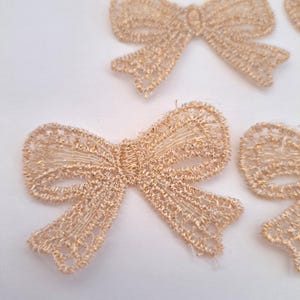 Op de afbeelding: Close-up van verschillende goudkleurige kanten strikken. De strikken zijn ingewikkeld ontworpen met een delicaat, open weefpatroon. De strikken zijn gerangschikt op een wit oppervlak, waarbij sommige elkaar overlappen. De goudkleur heeft een lichte glans.