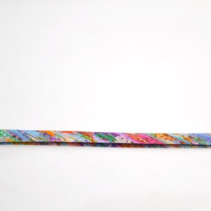 May include: A long, thin, flat strip of fabric featuring a vibrant, multi-coloured abstract pattern. The design includes blended streaks of orange, pink, blue, green, and yellow, creating a dynamic, painterly effect across the entire length of the ribbon.