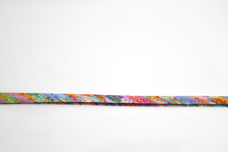 May include: A long, thin, flat strip of fabric featuring a vibrant, multi-coloured abstract pattern. The design includes blended streaks of orange, pink, blue, green, and yellow, creating a dynamic, painterly effect across the entire length of the ribbon.