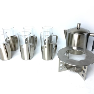 Op de afbeelding: Een roestvrijstalen theeset met zes heldere glazen mokken, elk met een zilveren handvat en houder. De set bevat een theepot, een warmhoudstandaard en een voet, allemaal in bijpassend roestvrij staal.