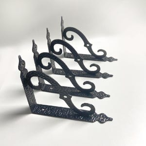 May include: Four black metal shelf brackets with a textured finish. Each bracket features a decorative scroll design and a pointed end. The brackets are arranged in a diagonal pattern against a white background.