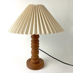 Op de afbeelding: Een houten tafellamp met een geplooide lampenkap van stof. De lampenkap is licht beige van kleur en de voet is van donkerbruin hout.