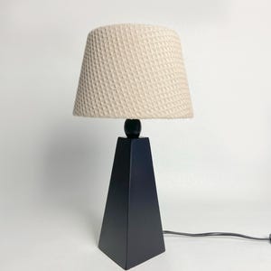 May include: A modern table lamp with a black, pyramid-shaped base and a beige, textured lampshade. The lamp features a black sphere connecting the base and the shade. The power cord is black.