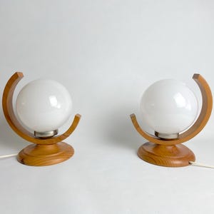 Op de afbeelding: Twee bijpassende tafellampen. Elke lamp heeft een witte bolvormige kap die wordt vastgehouden door een gebogen houten frame. De basis is ook van hout. De lampen zijn verbonden met een wit elektrisch snoer.