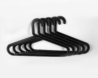 May include: A collection of six black plastic clothes hangers. Each hanger has a curved hook and a wide, flat body designed to support garments. The hangers are arranged in a slightly overlapping pattern against a white background.