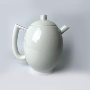 Op de afbeelding: Een witte theepot van keramiek met een rond lichaam en een tuit. De theepot heeft een eenvoudig, modern ontwerp.