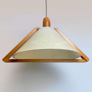 May include: A mid-century modern pendant light with a light cream-colored shade and a wooden frame. The shade has a textured appearance and is trimmed with a decorative edge. The light fixture is suspended from a gray cord and has a wooden top.