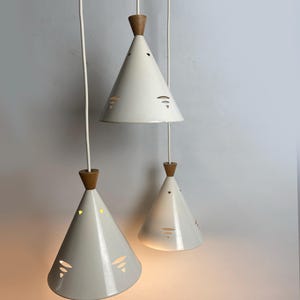 Op de afbeelding: Drie hanglampen met kegelvormige, crèmekleurige kappen en houten toppen. De kappen hebben uitgesneden patronen en hangen aan witte snoeren. De lampen hangen tegen een neutrale achtergrond.