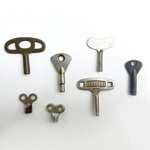 May include: A collection of seven vintage clock winding keys. The keys are made of metal and have different shapes and sizes. Some of the keys are silver, while others are brass or grey.
