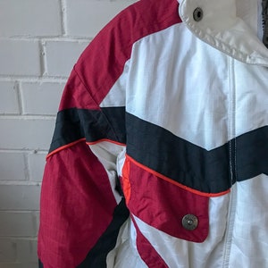 以下が含まれることがあります： 白い、赤と黒のスキーのジャケット。ジッパーで閉じて、ウエストにドローストリングが付いている。