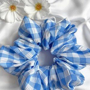 Könnte beinhalten: Ein großer blau-weiß karierter Scrunchie, ein Haaraccessoire aus Stoff, steht im Mittelpunkt des Bildes. Zwei weiße Blumen mit gelben Zentren befinden sich im Hintergrund. Der Scrunchie ist aus einem strukturierten Stoff gefertigt.