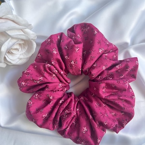 Puede incluir: Una gran goma para el pelo magenta con un estampado floral de pequeñas flores blancas y rosas. La goma para el pelo está hecha de tela fruncida y está colocada sobre un fondo de satén blanco. Una rosa blanca es visible en el fondo.