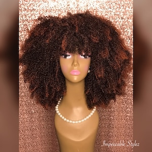 Puede incluir: Una peluca afro marr&oacute;n con flequillo en una cabeza de maniqu&iacute;. La peluca tiene un ligero tono marr&oacute;n rojizo.