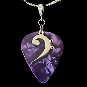 May include: A silver bass clef charm hangs from a silver chain on a purple guitar pick pendant. The pendant is displayed in a black velvet box.