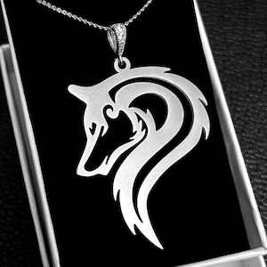 Op de afbeelding: Een zilverkleurige hangerketting met een wolfskopontwerp. De hanger wordt gepresenteerd in een open witte doos met een zwart interieur, tegen een zwarte, getextureerde achtergrond. De ketting heeft een fijne ketting.
