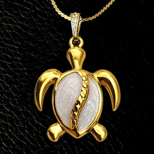 Pode incluir: Um colar com pingente de tartaruga em tom dourado, apresentando um detalhe de concha branca texturizada com uma crista central dourada. O pingente está suspenso por uma corrente dourada delicada e o passador possui pequenas pedras brilhantes.