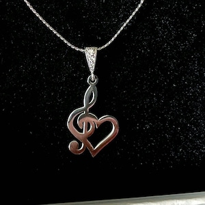 May include: A silver necklace featuring a treble clef charm shaped like a heart. The necklace is presented in a white box with a black velvet lining.