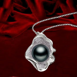 May include: A silver pendant necklace featuring a black pearl set within a shell-shaped design. The necklace is displayed against a red velvet backdrop.