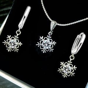 Op de afbeelding: Een zilveren set met een ketting en oorbellen in de vorm van een sneeuwvlok met een helder kristal in het midden van elke sneeuwvlok. De sieraden worden gepresenteerd in een zwarte fluwelen doos.