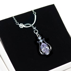 Może przedstawiać: Srebrny naszyjnik z wisiorkiem, prezentowany w białym pudełku prezentowym. Wisiorek ma kształt gruszki, jasnofioletowy kamień szlachetny osadzony w dekoracyjnej srebrnej ramie. Naszyjnik ma delikatny łańcuszek.
