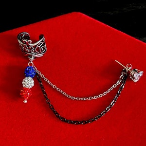 Op de afbeelding: Een zilveren oormanchet met een hangende ketting en drie kleine, gekleurde kralen. De ketting is verbonden met een zilveren oorbel met een heldere strasssteen.